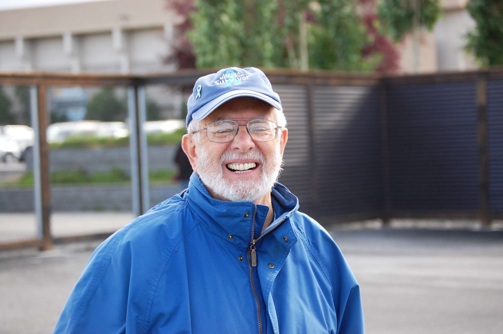 Photo of grey-haired man with a grey beard and glasses, wearing a blue jacket and baseball cap, smiling although the photographer has captured him with his eyes closed. The background is a blurred urban building.