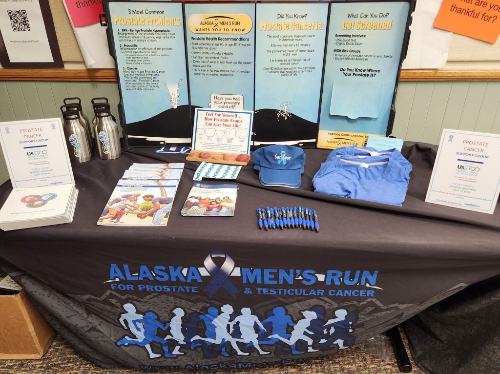 View forward and down of a table covered with a cloth printed with "Alaska Men's Run for prostate and testicular cancer" above artwork in blues representing a group of men running against a background of distant mountains. On the table are various brochures, pens, hats, t-shirts, water bottles, including posters promoting the UsToo support group. There is a backdrop of standing posters with information on prostate screening. 